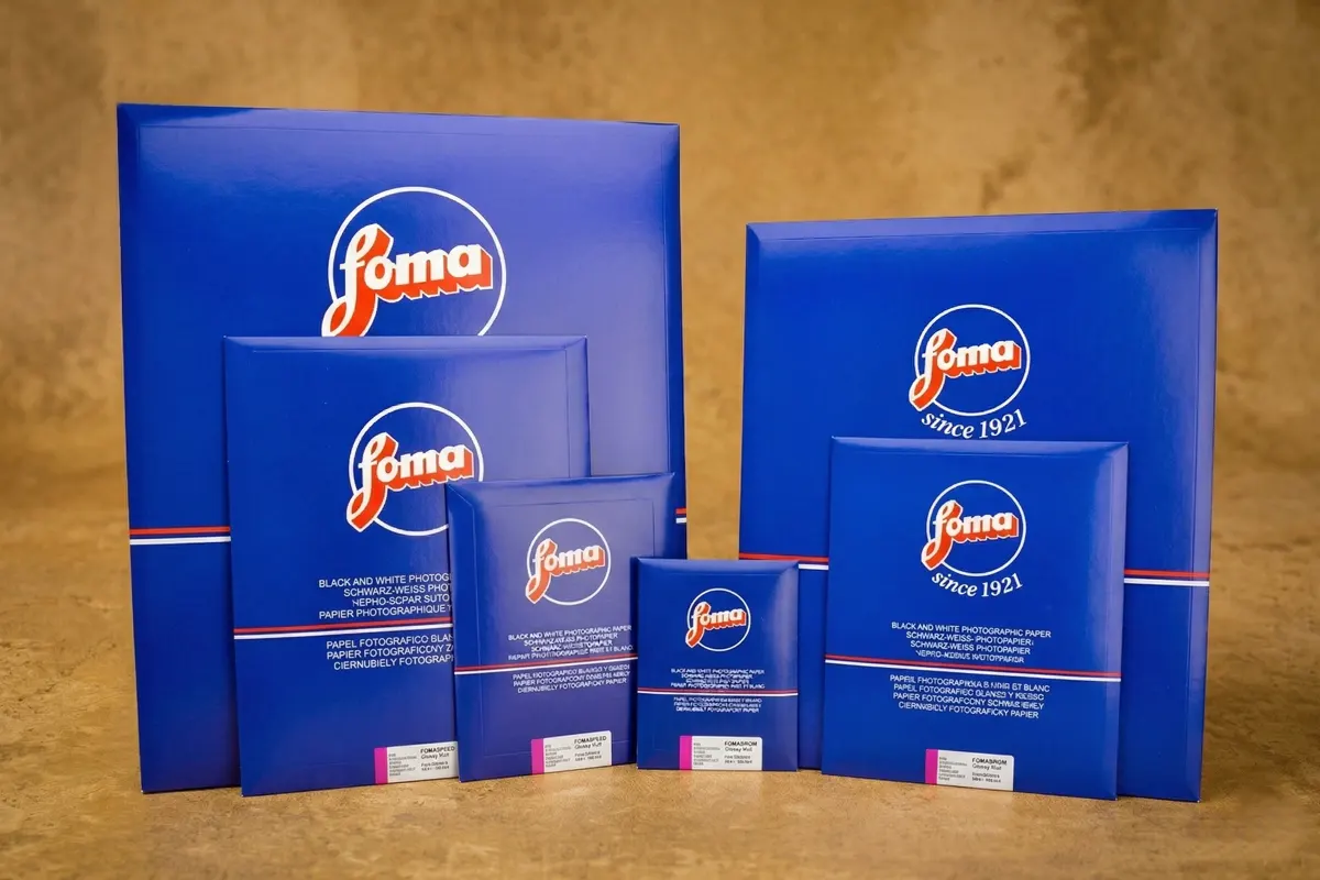 Sobres y cajas azules de papel fotográfico en blanco y negro de la marca Foma (Fomaspeed y Fomabrom) en diferentes tamaños sobre un fondo cálido.